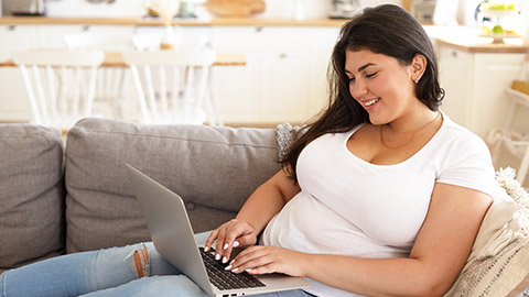 Side view of fashionable young overweight plump female freelancer sitting on sofa with portable computer on her lap