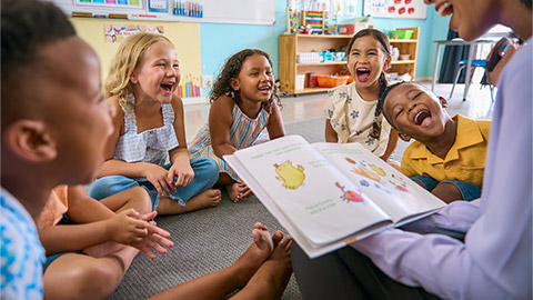 Female Primary Or Elementary School Teacher Reads Story To Multi-Cultural Class Seated In Classroom