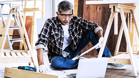 Elder man reading the computer while making wooden furniture