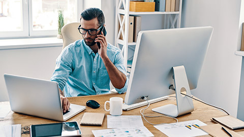 An accountant sitting at his desk talking on his phone while using his laptop