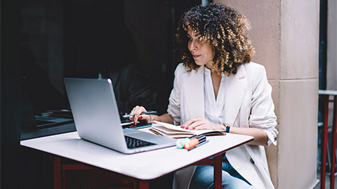 A person seated at a table outside, referencing their diary while working on a laptop