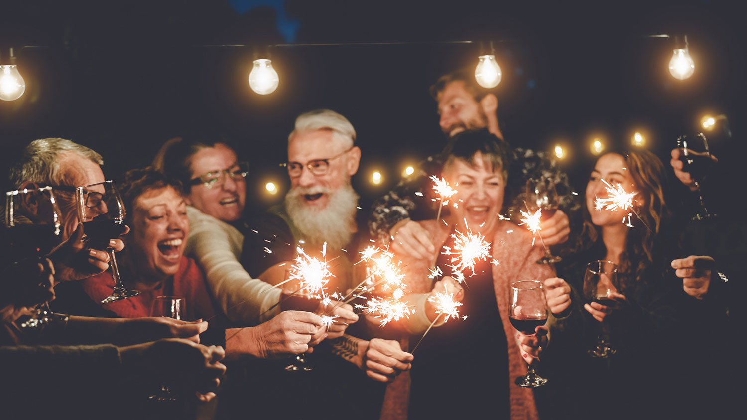 Happy family having at dinner party outdoor - Group of multiracial older and young people celebrating together drinking wine holding fireworks sparklers - Concept of youth and elderly parenthood