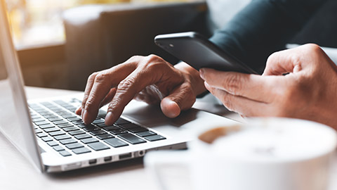 Close up image of hands typing on laptop while the other holds a mobile phone