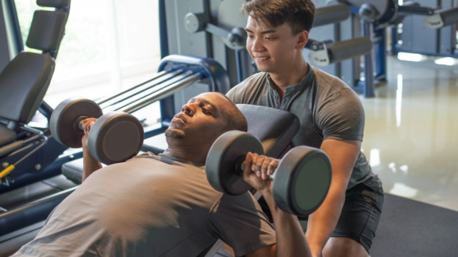 Male personal trainer spotting a client who is using dumb bells